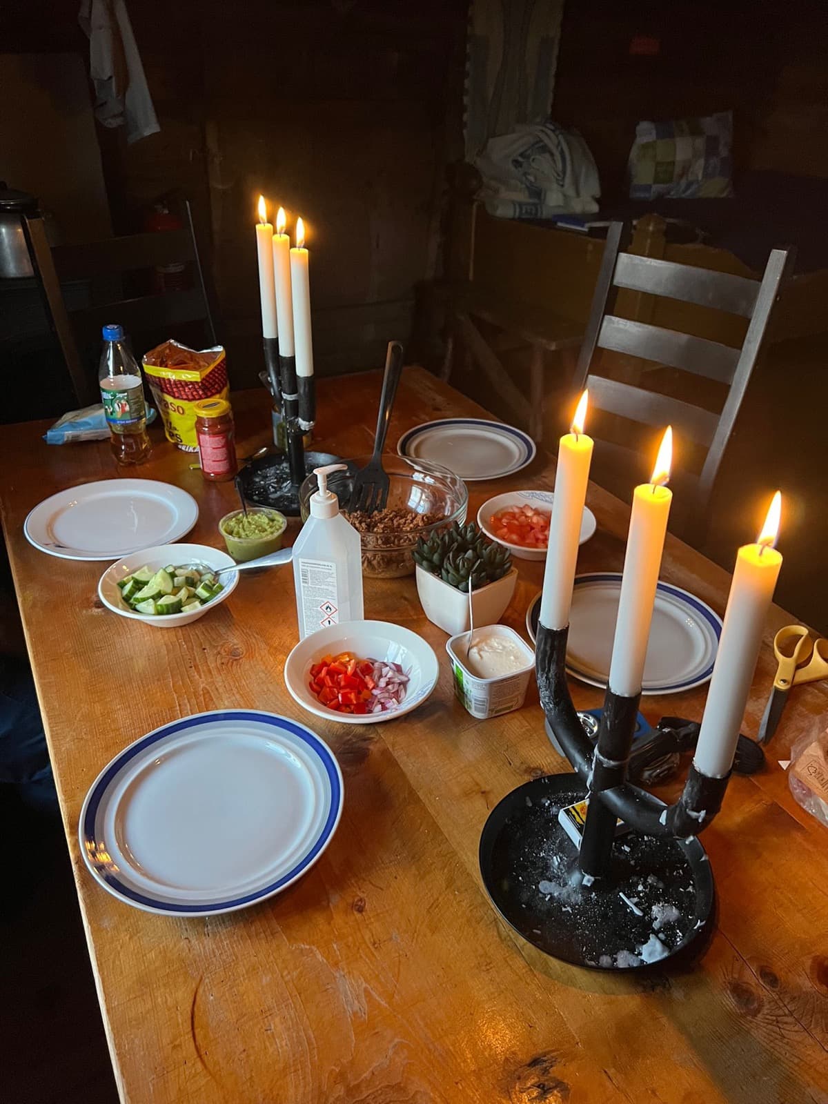 A wooden table lit by two candelabras, set with plates and bowls of taco toppings.
